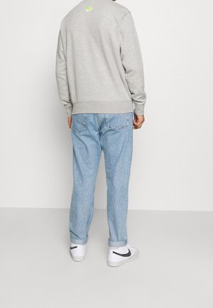 Mann trägt ein hellgraues Sweatshirt mit neonfarbigem Logo, hellblaue hochgekrempelte Jeans und weiße Turnschuhe mit schwarzem Logo, steht mit dem Rücken zur Kamera.