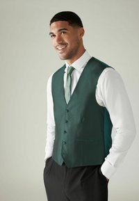 Dark green textured vest with five buttons, worn over a white shirt, and paired with a matching tie. Black trousers and subtle side pockets.