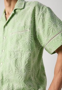 Short-sleeve, light green shirt made of textured fabric, featuring white sunburst embroidery, a chest pocket, and decorative open stitching.
