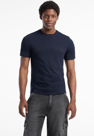 Marineblå kortærmet T-shirt lavet af bomuld med et tekstureret "GUESS JEANS" logo på brystet, parret med grå cargo bukser.