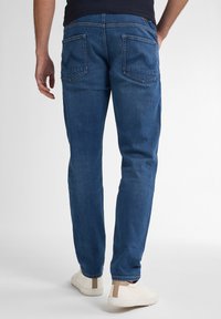 Vaqueros de mezclilla azul con corte recto, presentan dos bolsillos traseros y detalles de costura. Llevados con zapatillas blancas.