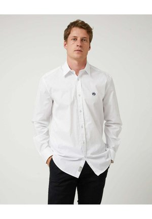 Hombre con cabello castaño claro que lleva una camisa blanca abotonada con un pequeño logo en el pecho izquierdo y pantalones negros, manos en los bolsillos, fondo neutro.