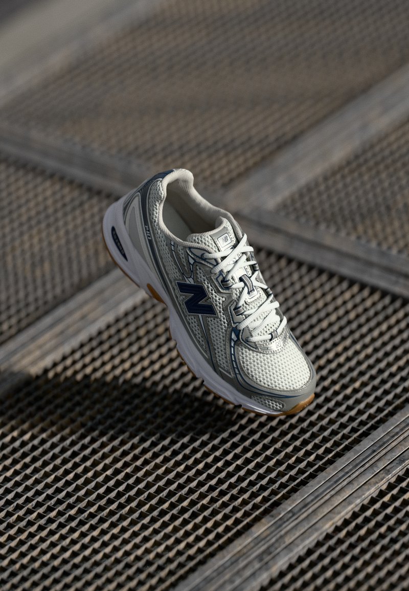Zapatilla New Balance blanca y gris con logo y cordones en azul marino sobre una superficie de rejilla metálica en luz natural.