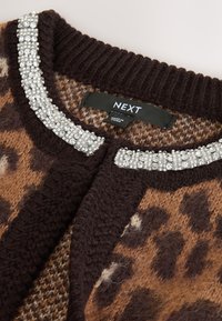 Ruskea leopardikuvioinen neuletakki, jossa on eläväpintainen neulos ja hopeiset helmiripustukset kaula-aukossa ja napituslistassa. Brändin merkintä näkyvissä.