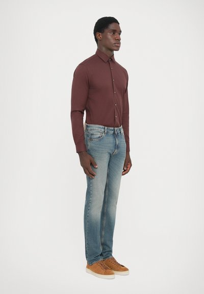 Mörkbrun långärmad skjorta, knäppt, i kombination med ljusblå jeans och beige sneakers. Modellen står i profil mot en enfärgad bakgrund.