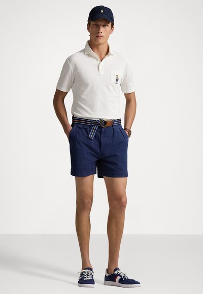 Hvid polo shirt med brystlomme, navy shorts med stribet bælte, navy baseball cap og navy sko med hvide accenter.