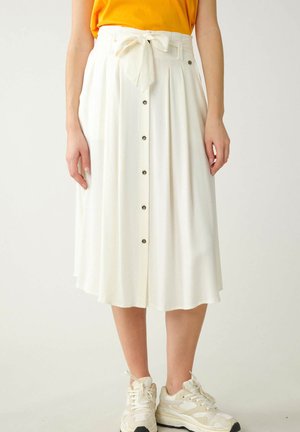 Jupe plissée blanche avec une ceinture à nouer, dotée d'une fermeture à boutons et d'une texture lisse. Associée à des baskets de couleur claire.