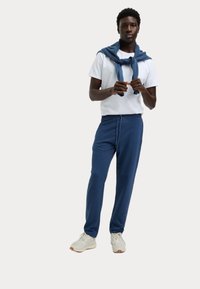 Marineblauwe joggerbroek met elastische tailleband en koord. Gecombineerd met een wit T-shirt en een blauwe sweater die over de schouders is gedrapeerd.