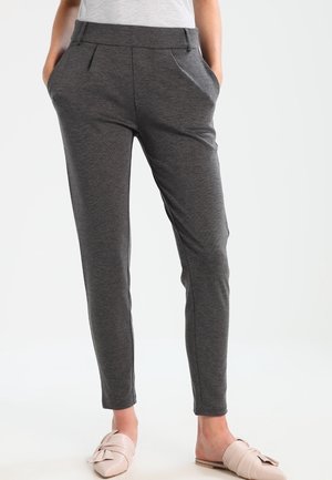 Nainen, jolla on päällään tummanharmaat slim fit -housut sivutaskuilla ja vaaleanpunaiset, liukuvat matalat kengät, seisoo valkoista taustaa vasten.