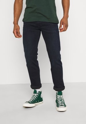 Homme portant un jean foncé retroussé, des baskets montantes vertes avec des lacets blancs et une chemise vert foncé, debout sur un sol uni.