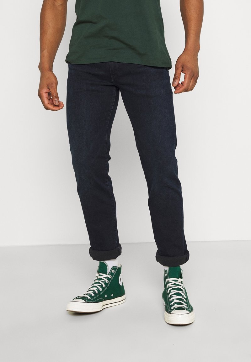 Homme portant un jean foncé retroussé, des baskets montantes vertes avec des lacets blancs et une chemise vert foncé, debout sur un sol uni.
