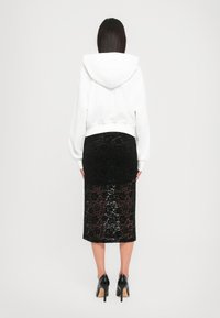 Donna in piedi di spalle, che indossa un hoodie bianco, una gonna midi nera in pizzo e scarpe con tacco alto nere, su uno sfondo semplice.
