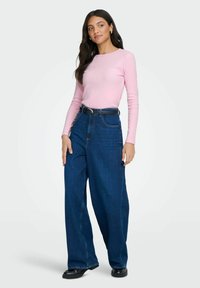 Lichtroze langmouwtop gecombineerd met hoge taille, wijde blauwe jeans, voorzien van een zwarte riem en glanzende zwarte schoenen.