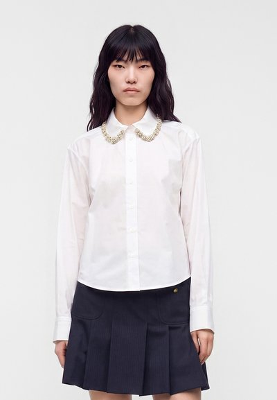 Femme portant une chemise blanche à boutons avec un col orné de perles et une jupe noire plissée, debout devant un fond clair uni.
