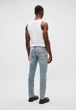 Mand iført lyseblå jeans, hvid ærmeløs skjorte og mørkebrune sko, står med ryggen til mod en ensfarvet baggrund.