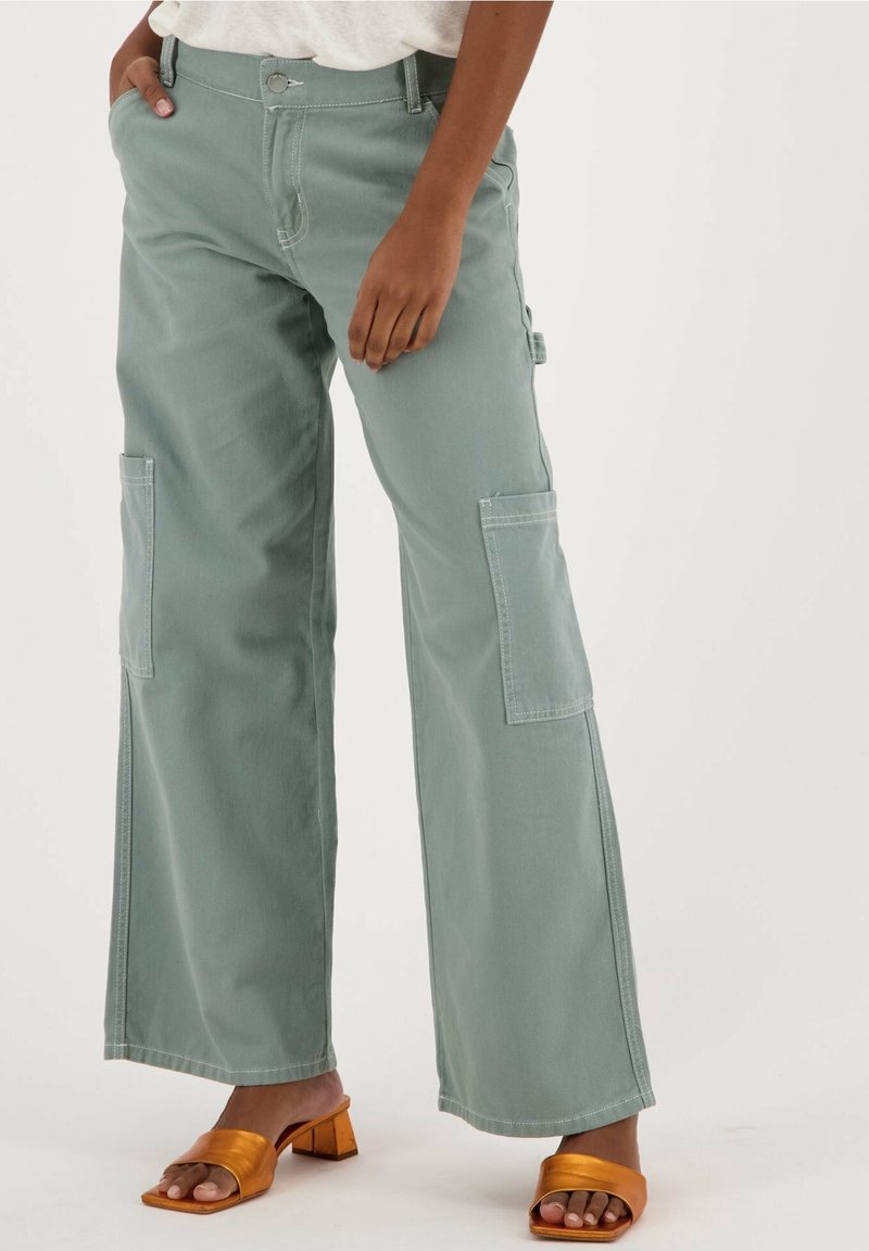 Femme portant un pantalon cargo large vert sauge clair avec poches avant et latérales, associé à des sandales à talons blocs orange à bouts ouverts.