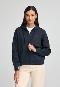 Felpa nera con zip, realizzata in misto cotone, con tasche laterali, cappuccio regolabile con cordino e un piccolo logo rosso sul petto sinistro.