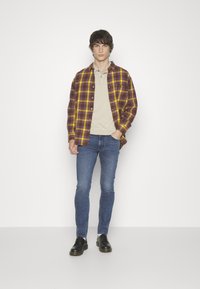 Camisa de franela a cuadros en púrpura, amarillo y marrón sobre una polo beige; jeans slim azul; zapatos negros. El modelo está de pie frente a un fondo blanco.
