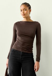 Nuori nainen, jolla on letitetyt hiukset, pukeutuneena tiukkaan ruskeaan pitkähiharaiseen puseroon ja mustiin housuihin, pitää ruskeaa käsilaukkua yksinkertaista taustaa vasten.