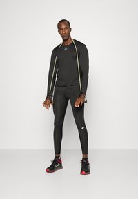Czarna, długorękawowa koszulka sportowa z odblaskowymi akcentami, zestawiona z czarnymi legginsami i czarno-czerwonymi butami do biegania. Widoczne logo Adidas.