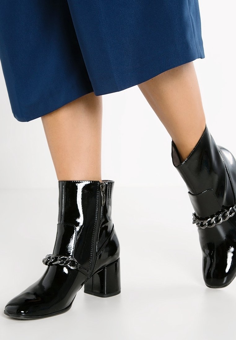 Bottines en cuir verni noir avec un talon épais et une chaîne décorative sur le haut. Détail de fermeture éclair sur le côté.