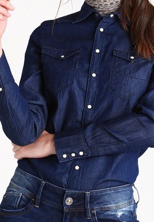 Frau trägt dunkelblaues Denimhemd mit weißen Druckknöpfen und blaue Jeans, posiert mit einem Arm verschränkt und dem anderen erhoben.