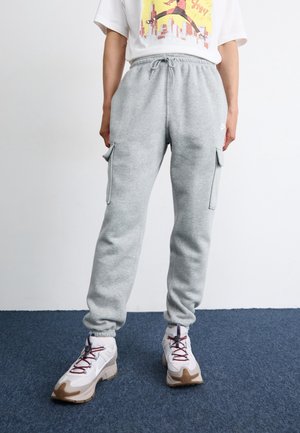 Person trägt hellgraue Cargo-Jogginghosen und weiße Sneakers und steht auf einem blauen Teppich vor einer weißen Wand.