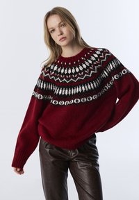 Pull en maille rouge avec un empiècement à motif noir, blanc et bordeaux. Coupe ample avec manches larges et poignets côtelés, associé à un pantalon en simili cuir noir.