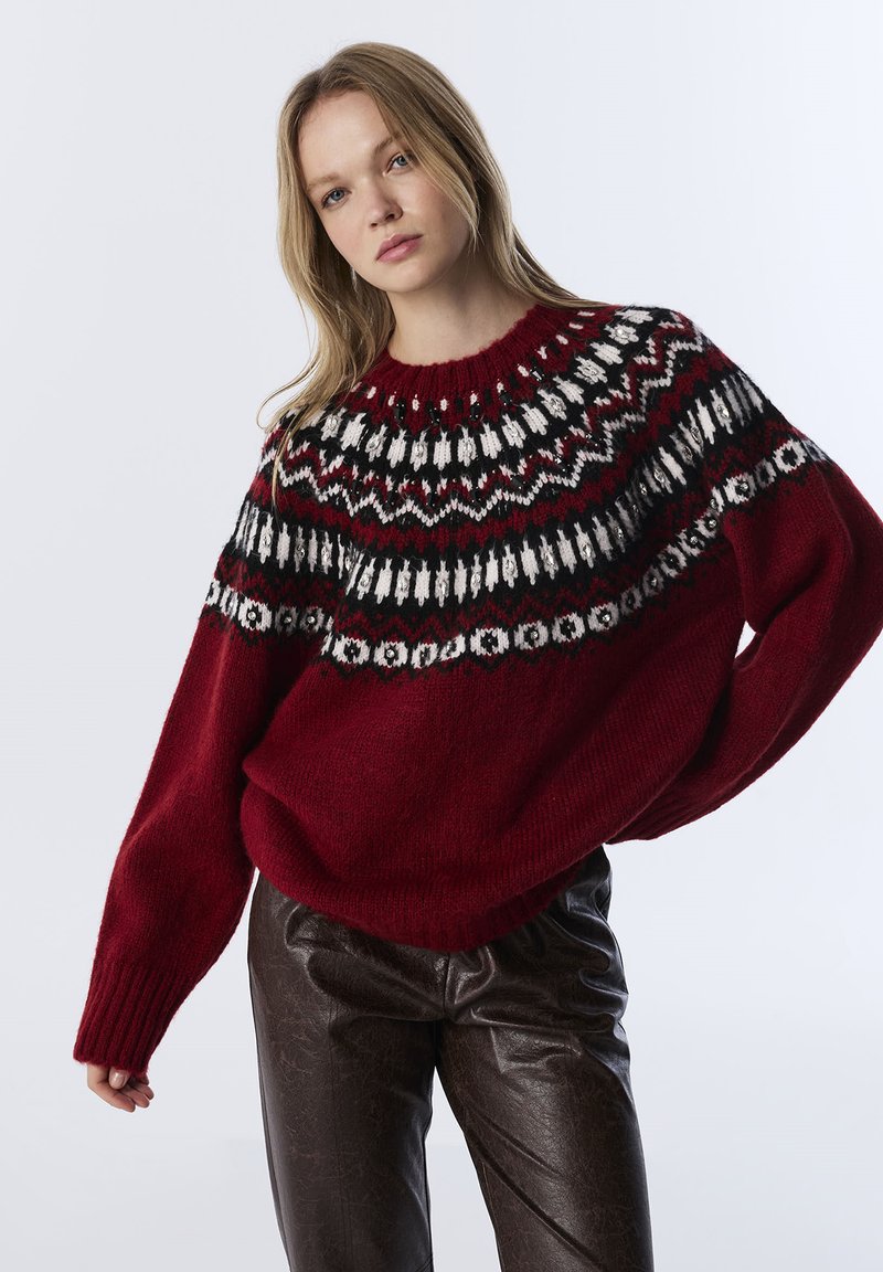 Pull en maille rouge avec un empiècement à motif noir, blanc et bordeaux. Coupe ample avec manches larges et poignets côtelés, associé à un pantalon en simili cuir noir.
