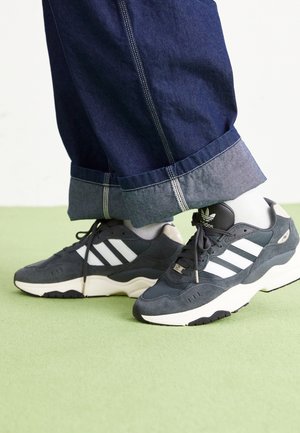 Tmavě šedé tenisky Adidas s bílými pruhy, nošené s ohrnutými tmavě modrými džíny a bílými ponožkami na světle zelené podlaze.