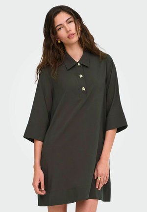 Femme portant une robe ample vert foncé avec trois boutons dorés et des manches larges, debout devant un fond clair uni.