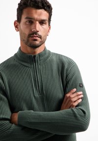 Groene, ribgebreide trui met een ritskraag en een logo patch op de linkermouw. Heeft een gestructureerde, zachte afwerking.