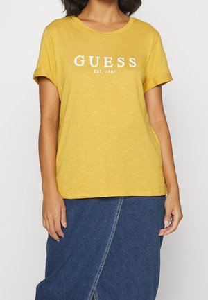 Osoba nosząca żółtą koszulkę Guess z napisem "GUESS EST. 1981" i niebieską dżinsową spódnicę z przednim rozcięciem.