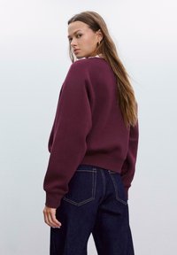 Burgundijska sweatshirt s spuštenim ramenima, rebrastim manžetama i rubom, u kombinaciji s tamnim trapericama koje imaju kontrastne šavove i stražnje džepove.