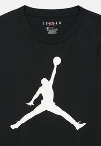 Musta puuvillainen T-paita, jossa on suuri valkoinen siluetti hyppäävästä hahmosta, joka pitää koripalloa. Pyöreä kaula-aukko ja normaali istuvuus.