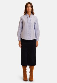 Lichtblauw gestreept shirt met ruffles aan de voorkant, knoopsluiting en hoge kraag, gecombineerd met een lange zwarte rok en bruine suède kniehoge laarzen.