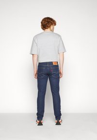 Camisa cinza de mangas curtas combinada com jeans azul escuro. Os jeans têm um corte clássico e detalhes de costura visíveis, com uma etiqueta de marca na parte de trás.