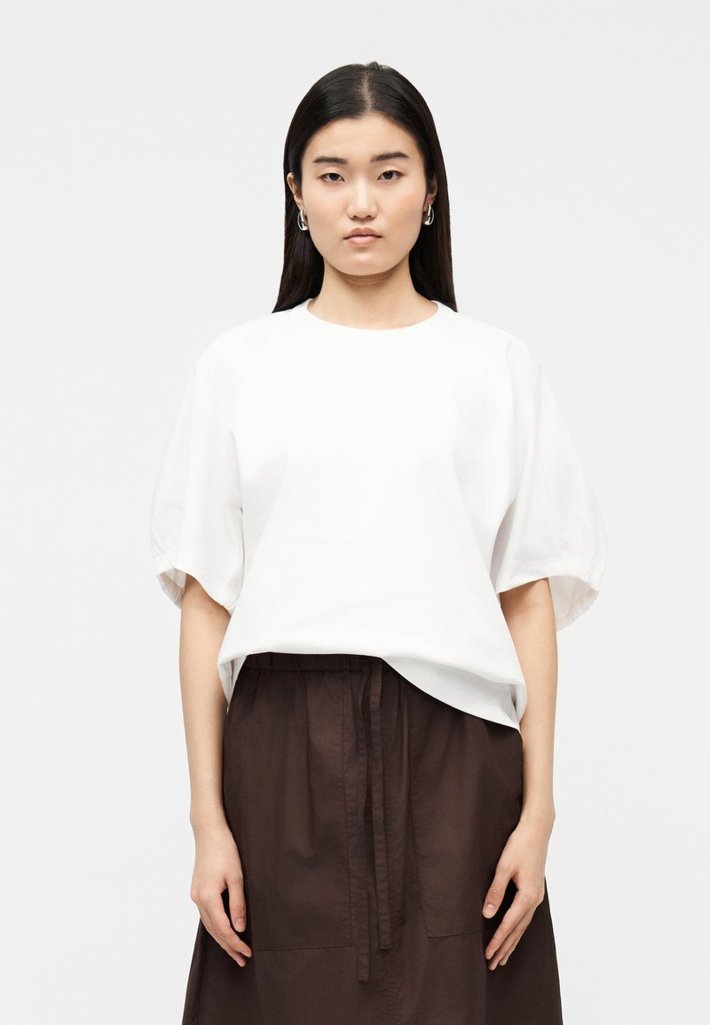 Νέα γυναίκα με μακριά σκούρα μαλλιά φοράει μια άνετη λευκή μπλούζα με κοντά μανίκια και σκούρο καφέ φούστα, στέκεται μπροστά σε απλό φόντο.