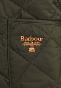 Quiltad olivgrön väst med en fickklaff och en broderad orange "Barbour"-logotyp med ett grafiskt designelement.