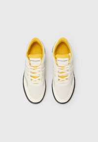 Witte sneaker met gele accenten en veters, voorzien van een geperforeerde textuur, drie witte strepen en een zwarte rubberen zool. Merkenlabel zichtbaar.