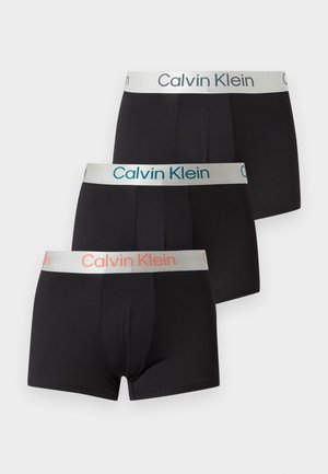 Młody mężczyzna ubrany w czarną bieliznę Calvin Klein, siedzący boso na ciemnej skórzanej sofie na jednolitym tle.