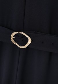 Tissu de robe noir à la texture lisse, présentant une large boucle dorée ondulée accentuant une ceinture. Le matériau semble léger et fluide.