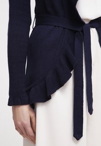 Námořnický kardigan s žebrovanou texturou, zdobený volánkovým lemem a páskem na zavázání v pase. Měkká tkanina s otevřeným předním designem.