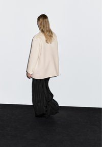Blazer oversize beige chiaro con texture liscia, abbinato a pantaloni neri larghi a pieghe con sottili righe verticali. Modella di spalle.
