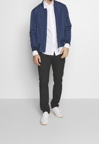 Muž oblečený v námořnicky modré bomber bundě, bílé košili na knoflíky, tmavě šedých kalhotách a bílých teniskách, stojící na pozadí bílé stěny.