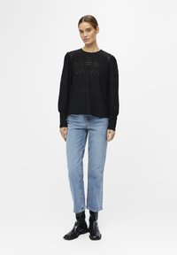 Pull noir en tricot avec un motif ouvert texturé, manches longues et col rond, associé à un jean droit bleu clair et des bottes noires.