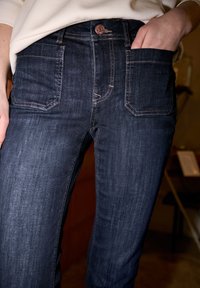 Personne portant un jean bleu foncé avec des poches avant, une main dans une poche, et un haut beige partiellement rentré.