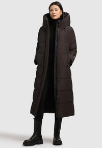 Long manteau d'hiver noir avec capuche, présentant un design matelassé, des fermetures éclair et à boutons-pression, ainsi que des poches latérales. Assorti à des bottes noires arrivant au genou.