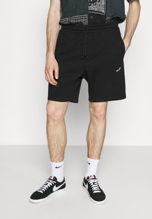 Homme portant un short noir Levi's, des chaussettes blanches Nike hautes et des baskets noires Nike, debout les mains près des poches devant un fond uni.