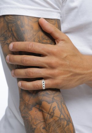 Hand mit silbernem Ring, die auf einem tätowierten Unterarm ruht, bekleidet mit einem weißen Kurzarmhemd.